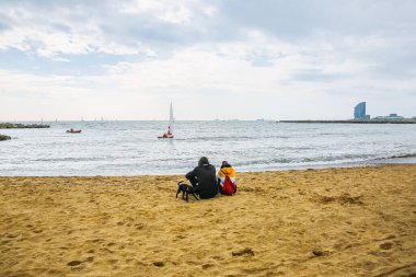 Çift sonbahar plajında oturuyor. Köpek kumda yürüyor. Atmosfer fotoğrafı. Barceloneta. Aşık bir adam ve kadın.