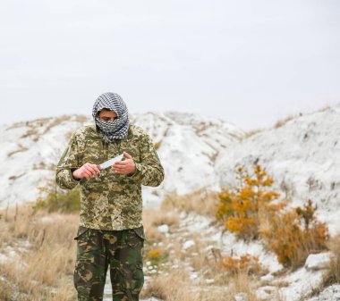 Asker kamuflaj üniforması içinde ve damalı keffiyeh shemagh bandanası içinde duruyor. Bıçaklı adam ıssız bir yerde, dışarıda..