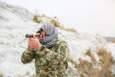 Asker kamuflaj üniforması içinde ve damalı keffiyeh shemagh bandanası içinde duruyor. Ellerinde dürbün var. İnsan ıssız bir yerde, açık havada..