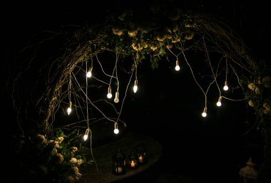 Düğün töreninde beyaz çiçek kemeri. Açık hava dekorasyonu. Romantik köy tarzı. Akşamları lambalar.