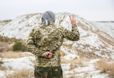 Soldier standing in camouflage uniform and checkered keffiyeh shemagh bandana. Man with knife behind back in the abandoned deserted place. Betraying of enemies. No war and peaceful sign.