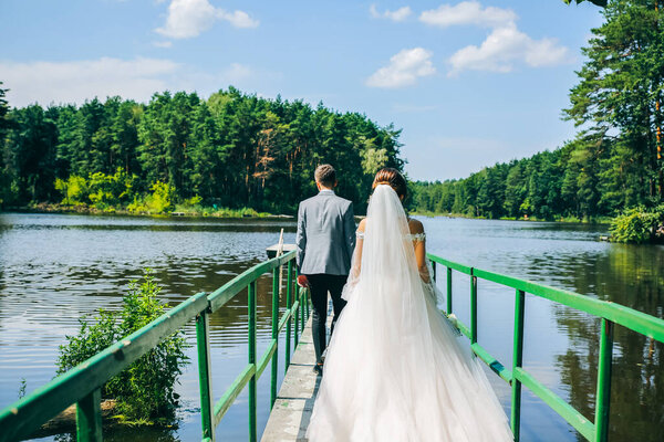Bride and groom are walking on the bridge. Wedding day photo near the lake and forest. Love story. Beautiful long sleeve dress. Lace veil.