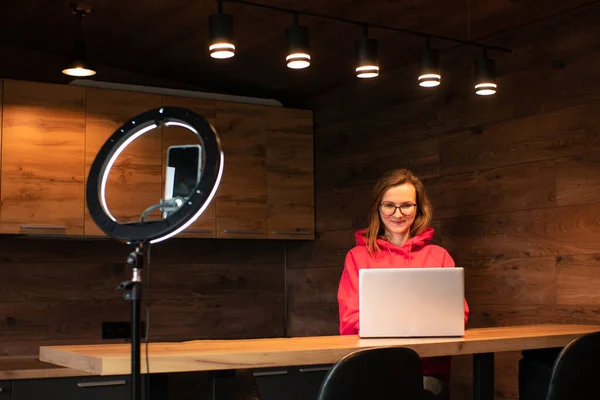 Social media influencer creating content at modern studio sharing online stream using smartphone and laptop. Girl blogger smiling and speaking recording video  with ring lamp. Online training courses.