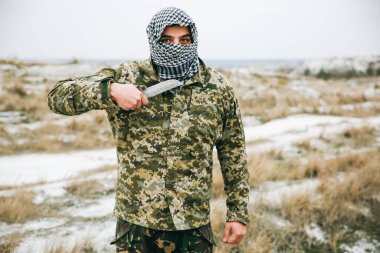 Asker kamuflaj üniforması içinde ve damalı keffiyeh shemagh bandanası içinde duruyor. Bıçaklı adam ıssız bir yerde, dışarıda..