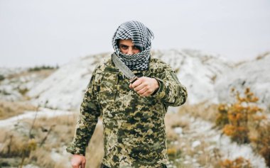 Asker kamuflaj üniforması içinde ve damalı keffiyeh shemagh bandanası içinde duruyor. Bıçaklı adam ıssız bir yerde, dışarıda..