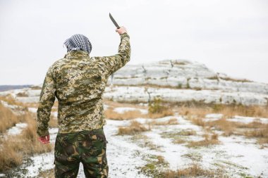 Asker kamuflaj üniforması içinde ve damalı keffiyeh shemagh bandanası içinde duruyor. Bıçaklı adam ıssız bir yerde, dışarıda..