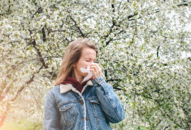 Çiçeklerle dolu bir ağacın yakınındaki mendile hapşıran bir kadın. Mevsimlik bahar alerjisi var..