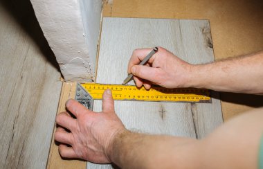 İşçi dairede döşeme kaplaması yapıyor. Mezurayı ve kalemi elinde tut. Onarım onarımı. Ahşap parke kalasları içeride..