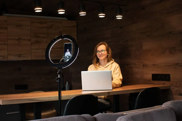 Social media influencer creating content at modern studio sharing online stream using smartphone and laptop. Girl blogger smiling and speaking recording video  with ring lamp. Online training courses.