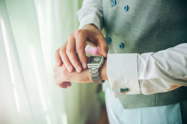 Açık gri takım elbiseli damat pencerenin yanında duruyor. Adam saatine bakıyor. Güzel kıyafetler..