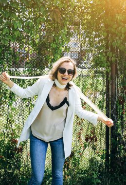 Havalı hippi kız dışarıda sarmaşık yapraklarının yanında duruyor. Beyaz ceketli ve kemerli genç kadınlar için modaya uygun bir fotoğraf. Sarışın, kısa saçlı ve gözlüklü. Yaz yaşam tarzı fotoshoot.