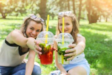 İki hippi kız gülüyor ve dışarıda yeşil çimlerin üzerinde kokteyl içiyorlar. Buzlu alkolsüz soğuk içecekler. Mojito ve çilekli limonata. Tatiller için mutlu bir yaşam tarzı.