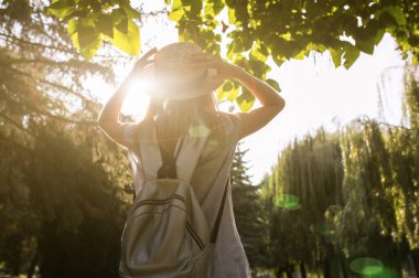 Hasır şapkalı şık hippi kız dışarıda bekliyor. Mavi elbiseli kadın. Gümüş gri sırt çantası. Seyahat tatili konsepti. Yaz yaşam tarzı fotoğrafı. Güneşin ve ağaçların gölgeleri.