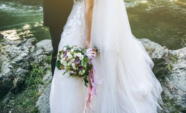 Düğün çifti dağ nehrinin yanında kucaklaşıyor. Damat ve gelin ellerinde buketle. Güzel çiçekler. Doğa Şelalesi.