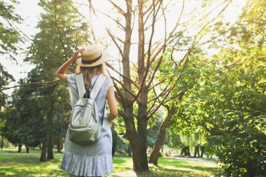Hasır şapkalı şık hippi kız dışarıda bekliyor. Mavi elbiseli kadın. Gümüş gri sırt çantası. Seyahat tatili konsepti. Yaz yaşam tarzı fotoğrafı. Güneşin ve ağaçların gölgeleri.