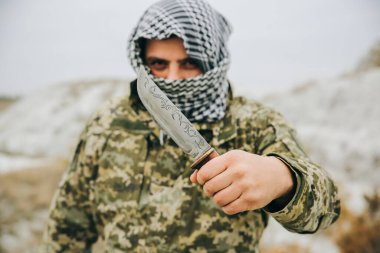 Asker kamuflaj üniforması içinde ve damalı keffiyeh shemagh bandanası içinde duruyor. Terk edilmiş bir yerde bıçaklı bir terörist var. Savaş konsepti.