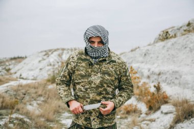 Asker kamuflaj üniforması içinde ve damalı keffiyeh shemagh bandanası içinde duruyor. Terk edilmiş bir yerde bıçaklı bir terörist var. Savaş konsepti.