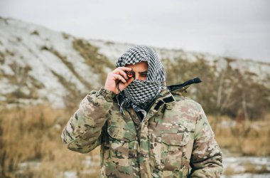 Asker kamuflaj üniforması içinde ve damalı keffiyeh shemagh bandanası içinde duruyor. Ellerinde dürbün var. Terörist terk edilmiş bir yerde, dışarıda. Savaş konsepti.