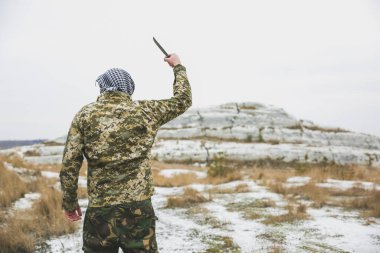 Asker kamuflaj üniforması içinde ve damalı keffiyeh shemagh bandanası içinde duruyor. Terk edilmiş bir yerde bıçaklı bir terörist var. Savaş konsepti.