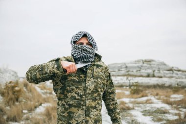Asker kamuflaj üniforması içinde ve damalı keffiyeh shemagh bandanası içinde duruyor. Terk edilmiş bir yerde bıçaklı bir terörist var. Savaş konsepti.