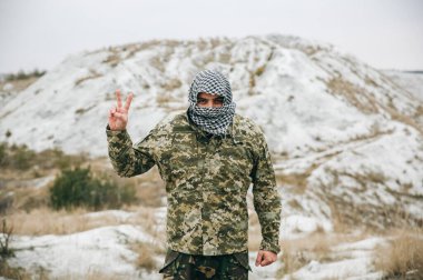 Asker kamuflaj üniforması içinde ve damalı keffiyeh shemagh bandanası içinde duruyor. Terörist terk edilmiş bir yerde, dışarıda. Savaş kavramı ve barışçıl işaret yok.