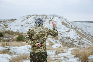 Kamuflaj üniforması ve damalı keffiyeh shemagh bandana giymiş bir asker. Terk edilmiş bir yerde sırtında bıçakla terörist. Düşmanlarına ihanet etmek. Savaş yok ve barış işareti yok..