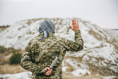 Kamuflaj üniforması ve damalı keffiyeh shemagh bandana giymiş bir asker. Terk edilmiş bir yerde sırtında bıçakla terörist. Düşmanlarına ihanet etmek. Savaş yok ve barış işareti yok..