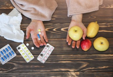 Grip olmuş hasta bir kız, tahta masada elinde haplar ve meyvelerle oturuyor. Soğuk algınlığı olan kadın ilaç ve elmaları elinde tutuyor. Doğal vitaminler ya da yapay konsept. Sağlık konsepti seçimi.