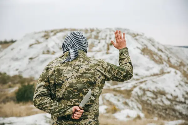 Kamuflaj üniforması ve damalı keffiyeh shemagh bandana giymiş bir asker. Terk edilmiş bir yerde sırtında bıçakla terörist. Düşmanlarına ihanet etmek. Savaş yok ve barış işareti yok..