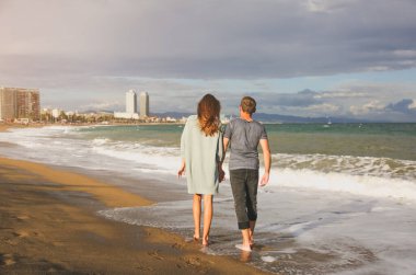 Güneşli bir günde kumsalda yürüyen aşık bir çift. Denizdeki dalgaların yanında şık hipsterler. Erkek ve kadın sarılıp öpüşüyor. Balayı. Tatil seyahati konsepti. Barselona İspanya görüşleri.
