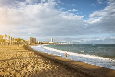 Güneşli bir günde kumsalda çanta taşıyan bir kız. Denizdeki dalgaların yanında şık bir hipster. Tatil seyahati konsepti. Barselona İspanya.