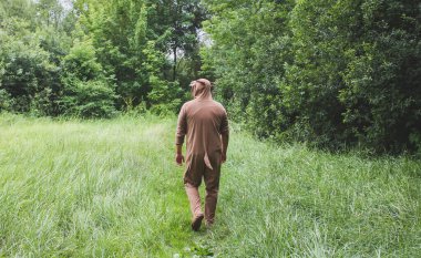 Genç adam ormanda bir inek kostümü içinde yürüyor. Komik hayvan pijamalı adam doğada uyku tulumu giyiyor. Parti için Cadılar Bayramı fikirleri.