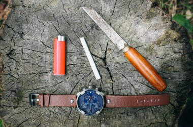 Bid elegant men's watch, old rusty knife, cigarette and lightener are lying on the tree truck in the forest. Accessories for stylish fashion background.