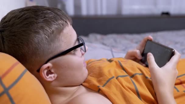 Nastolatek Okularach Gra Gry Wideo Smartfonie Leżąc Przytulnym Miękkim Łóżku — Wideo stockowe