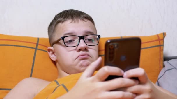 眼鏡の少年は 毛布の下にベッドに横たわっています 画面上で見て グライマスを作る 子供の頭を振る 笑みを浮かべて 面白いスマートフォン上でオンラインチャット 10代の若者が寝室の枕の上で休んでいる — ストック動画