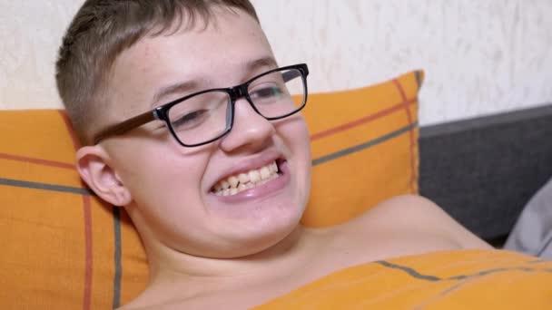 Ένας Έφηβος Γυαλιά Ξαπλωμένος Στο Κρεβάτι Κάνει Πρόσωπα Δείχνει Δόντια — Αρχείο Βίντεο