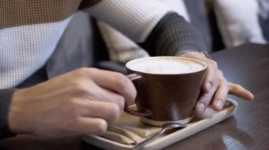 Kahve fincanı kapuçino içen adam, kafedeki masada köpüklü latte. Eller yukarı. Kahve dükkanında kremalı sıcak kahvenin tadını çıkar. Memnun oldum. Aroma. Öğle yemeği vakti. Hizmet. Yaşam tarzı. Rahatlama. Müşteri.