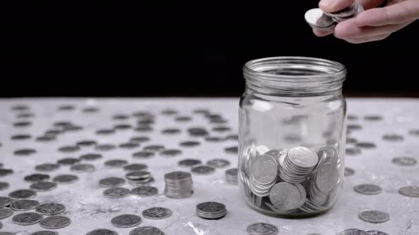 手把一捧累积的银硬币放进玻璃瓶里 小猪银行 一堆散落的乌克兰美分 硬币在桌子上 黑色背景 金融危机 预算节约 — 图库视频影像