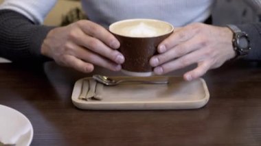 Kahve fincanı kapuçino içen adam, kafedeki masada köpüklü latte. Eller yukarı. Kahve dükkanında kremalı sıcak kahvenin tadını çıkar. Memnun oldum. Aroma. Öğle yemeği vakti. Hizmet. Yaşam tarzı. Rahatlama. Müşteri.