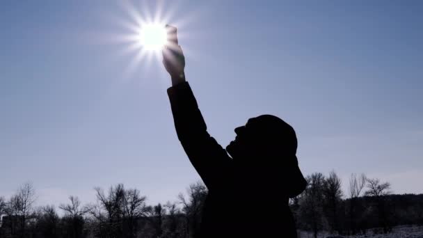 男性は冬の森で携帯電話の信号をキャッチしようとしています シルエットの男は インターネット 接続を求めて明るい太陽 青い空に手を伸ばします ホライズン ライフスタイル — ストック動画