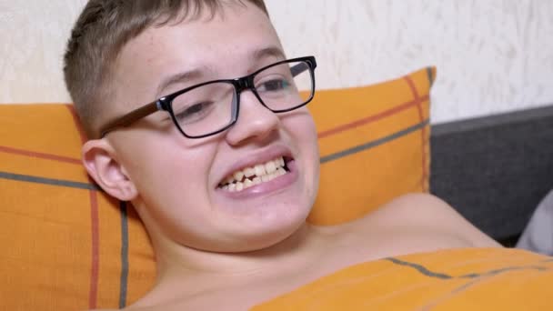 一个带着眼镜躺在床上的青少年 做鬼脸 露出牙齿 鬼鬼祟祟的 累了的男孩休息 睡觉前到处鬼混 改变了面部表情 玩得很开心 带着牙齿的宽笑 — 图库视频影像