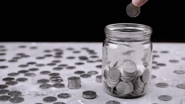 手把一盒五美分的乌克兰硬币扔进装满硬币的玻璃瓶里 桌子上有一堆散落的硬币 小猪银行 黑色背景 预算节约 — 图库视频影像