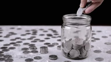 Bir avuç dolusu birikmiş gümüş parayı cam kavanoza koyan eller. Domuz kumbarası. Dağınık Ukrayna sentleri, masaya bozuk paralar. Siyah arka plan. Finansal kriz, bütçe tasarrufu. Yoksulluk.