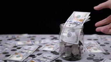 Kadın El, Bozukluklarla dolu bir kavanozdan 100 Dolarlık banknotlar alıyor. Bir yığın bozuk para ve banknotlar masanın üzerinde. Hisse. Domuz kumbarası. Harcama, bütçe, tasarruf, para birikimi.