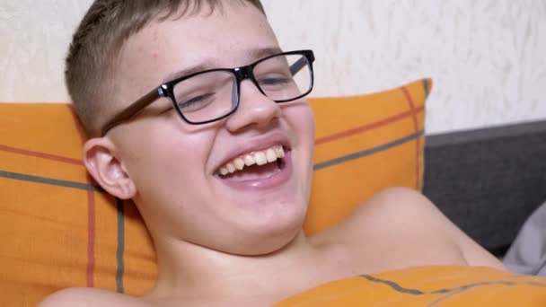 メガネの中の笑顔の幸せな10代がベッドの上に横たわっています 陽気な少年はベッドに行く前にリラックスして 面白い映画を見てください 子供の歯を持つ広い笑顔 子供の感情 — ストック動画