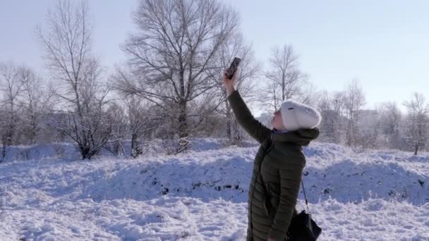 微笑的女孩正试图在冬天的森林里抓住手机的信号 女人伸出手智能手机蓝天寻找一个连接 — 图库视频影像