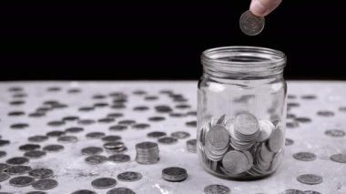 Hand Throws One Five-Kopeck Ukrainian Coin into a Glass Jar Filled with Coins. A Pile of scattered coins is on table. Stock. Piggy bank. Black background. Financial crisis, budget savings. Poverty.