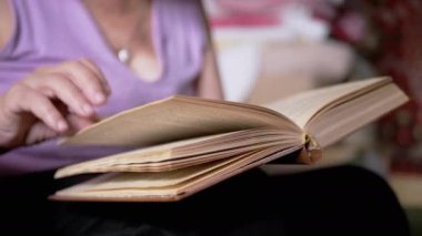 Kitap okuyan yaşlı bir kadın, Kapalı Kapılar 'daki güneş ışınlarında sayfaları çeviriyor. Yakın plan. Bir emeklinin elinde açık bir kitap vardır, çalışır ve emekliliğinde evde dinlenir. Yaşlıların yaşam tarzı..