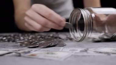 Cam kavanozu gümüş paralarla dolduran bir kadın. Beton zeminde Ukrayna sentleri ve 100 dolarlık banknotlar yatıyor. Bütçe, ödeme, harcamalar, tasarruflar. Finansal kriz. Nakit.