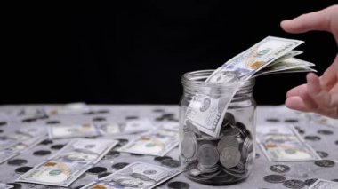 Kadın El, Bozukluklarla dolu bir kavanozdan 100 Dolarlık banknotlar alıyor. Bir yığın bozuk para ve banknotlar masanın üzerinde. Hisse. Domuz kumbarası. Harcama, bütçe, tasarruf, para birikimi.